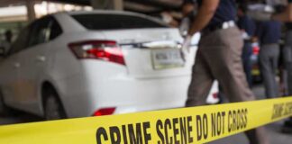 Yellow crime scene tape in front of a white car with investigators nearby